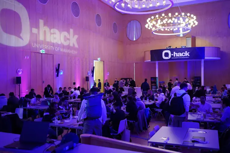 Participants at their tables in the university hall, the Q-Hack logo projected on the wall